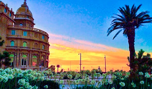 Imagem de um belo pôr do sol na cidade com árvore de palmeiras, edifícios históricos e jardim florido, transmitindo tranquilidade e beleza natural.