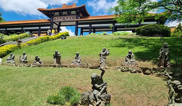 Vista do templo oriental, com estátuas em pedra de figuras em um jardim verdejo e céu azul ao fundo, destacando elementos da cultura asiática.