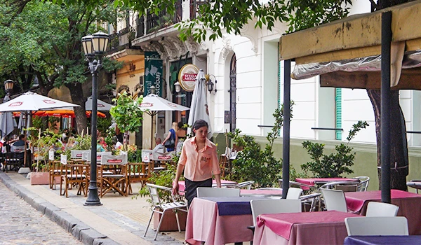 Rua charmosa com mesas de restaurante ao ar livre, cadeiras e guarda-sóis, em um ambiente urbano com árvores e iluminação aconchegante. Viagem para Buenos Aires.