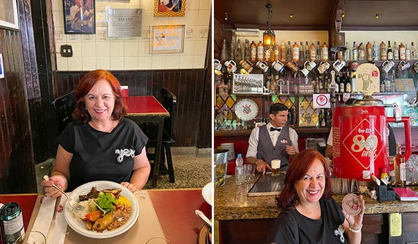 Deusa Rodrigues na frente de uma mesa com comida em um restaurante brasileiro, ambiente acolhedor decorado com garrafas e vinho ao fundo.