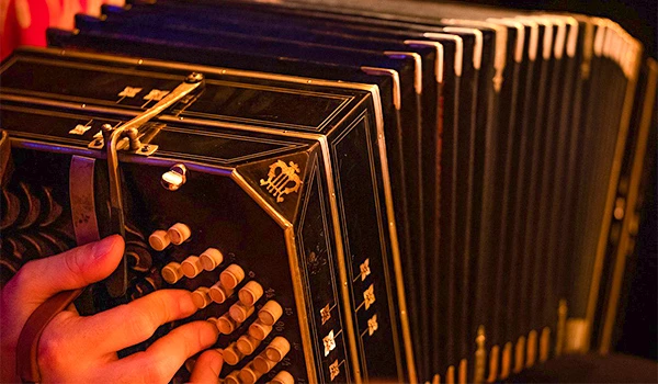 Imagem de um acordeon preto em close, com detalhes em botões e teclas visíveis, sendo manuseado por uma pessoa sob iluminação quente. Ideal para fãs de instrumentos musicais ou artistas de música ao vivo.