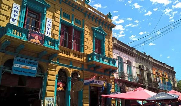 Foto de uma rua colorida com edifícios históricos em tons vibrantes de amarelo, azul e rosa, uma placa de artesanato e uma cobertura de guarda-sol rosada. O céu azul com algumas nuvens indica um dia ensolarado na cidade.