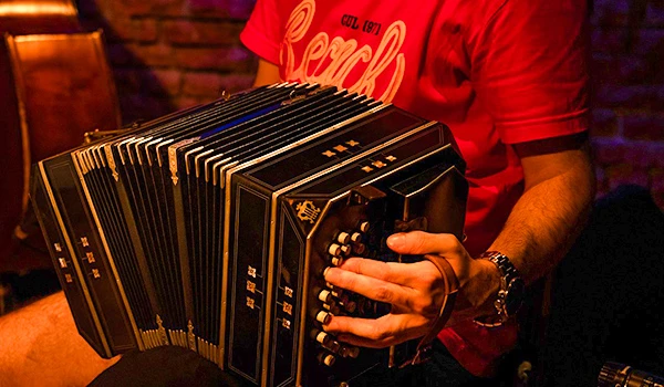Pessoa tocando acordeon em ambiente descontraído, usando camiseta vermelha com estampa "Beach" e relógio no braço esquerdo.