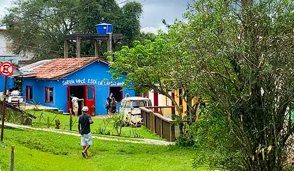 Imagem de uma casa colorida com a frase 'Sorria, você está entre amigos' em um ambiente rural. Pessoas estão na entrada, rodeadas por natureza. Vilarejo de Lavras Novas.