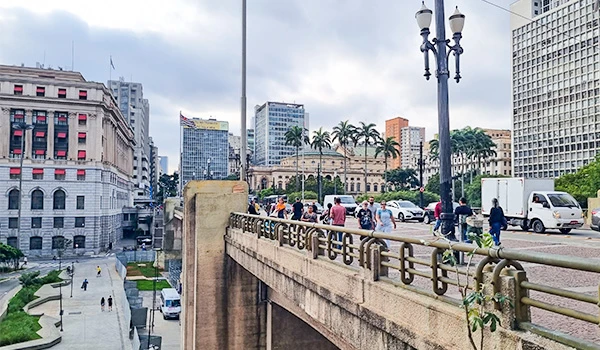 Vista do Viaduto do Chá com prédios altos, veículos e pedestres, em frente a um muro de concreto com guarda-corpo e poste de iluminação, com palmeiras ao fundo e céu nublado.