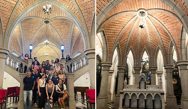 Foto de interior da  Cripta da Catedral da Sé, com arquitetura de tijolos aparentes e arcos altos, duas perspectivas mostrando corredores e nave com iluminação e vitrais, com grupo de pessoas em primeiro plano.