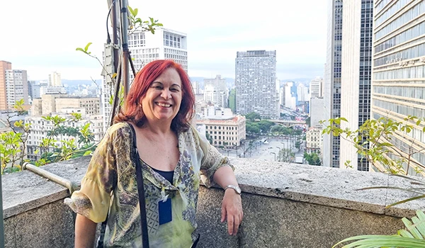 Deusa Rodrigues sorrindo em um mirante ao ar livre entre arranha-céus, usando vestido estampado e acessórios, com vista urbana ao fundo. Terraço do prédio da Prefeitura de São Paulo.