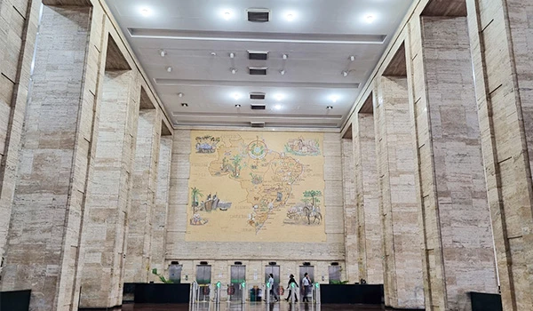 Hall interno com paredes revestidas de pedra, teto com luminárias embutidas e um grande mapa antigo na parede ao fundo, com mesas e objetos expositivos. Sede da Prefeitura de São Paulo.