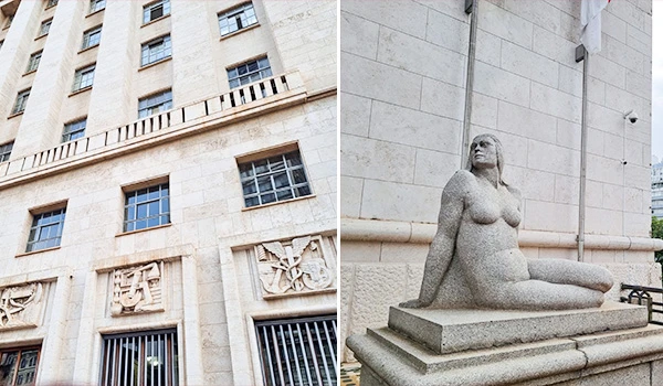 Detalhes arquitetônicos de fachada de prédio com fachada de pedra e janelas retangulares, ao lado de uma estátua de mulher clássica de pedra em um pedestal, em ambiente urbano. Sede da Prefeitura de São Paulo.