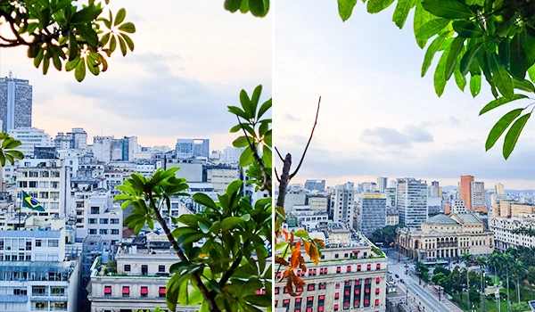 Vista panorâmica de uma cidade com prédios e ruas urbanas, enquadrada por folhas verdes de árvores em primeiro plano, com céu nublado ao fundo.