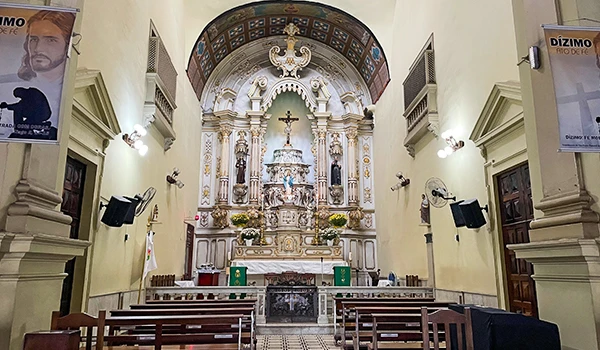Imagem do interior de uma igreja católica com altar decorado, cruz, estátuas santas, e bancos de madeira, ideal para orações e missas.