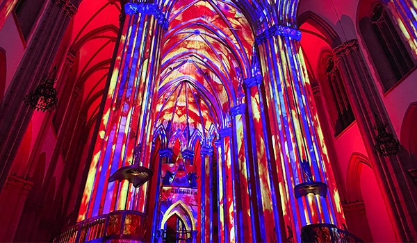 Interior de igreja iluminada com projeções de cores vibrantes em paredes e colunas, criando um ambiente artístico e iluminado.