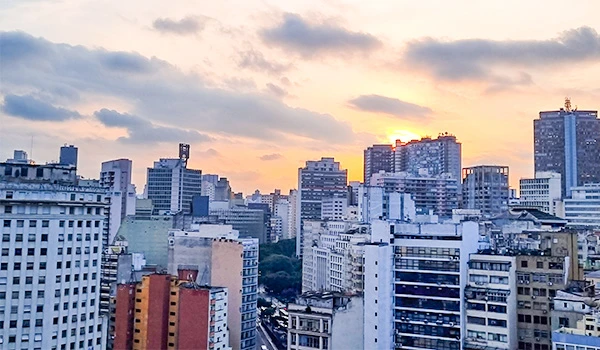 Vista panorâmica de uma cidade com diversos prédios residenciais e comerciais ao entardecer, céu com nuvens e luz dourada se espalhando entre os edifícios, em perspectiva urbana urbana.