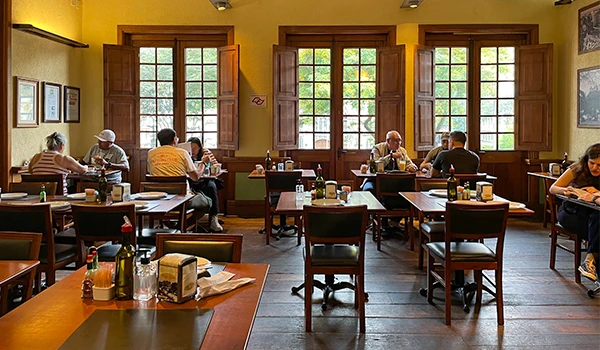 Imagem de interior de restaurante com clientes sentados às mesas, janelas de madeira abertas, ambiente aconchegante e decorado com quadro na parede.
