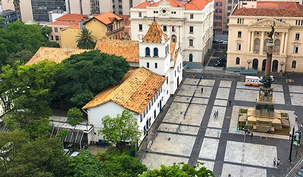 Vista aérea do centro histórico do Páteo do Colégio no entorno outros prédios clássicos de telhados laranja e áreas verdes, destacando a praça pavimentada e monumentos.