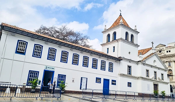 Fachada de prédio histórico branco com janelas azuis e porta azul, em estilo colonial, com torre sineira e telhado de cerâmica ao fundo sob céu parcialmente nublado. Páteo do Colégio.