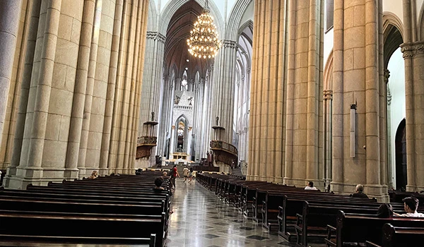 Interior de uma catedral com colunas altas de pedra dourada, arcos em estilo neogótico, bancos e um lustre ao fundo, vista central do altar. Cripta da Catedral da Sé.