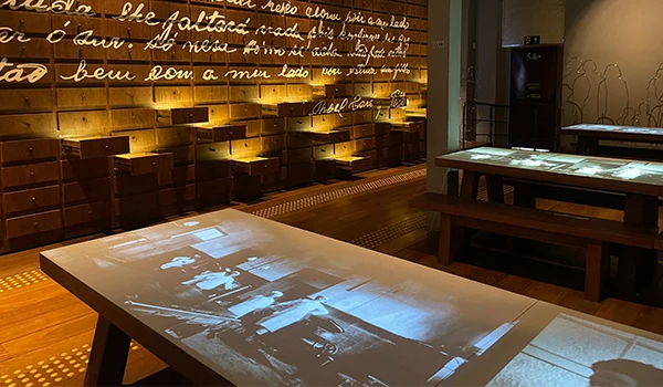 Imagem de um ambiente de exposição com mesas interativas que projetam fotografias históricas e um fundo de paredes com textos iluminados. A cena é rica em detalhes que remetem à memória cultural. Ideal para verificar a ancestralidade no Museu da Imigração.