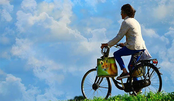 Pessoa pedalando uma bicicleta sob céu azul com nuvens, usando roupa branca e carregando uma bolsa com cachorro, representando liberdade e conexão com a natureza.
