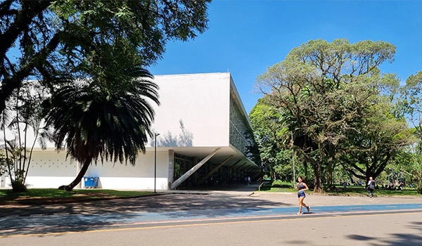 Imagem de um dos prédios do Parque Ibirapuera, rodeado por árvores verdes, sob um céu azul claro.