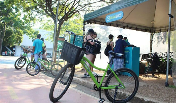 Pessoas utilizando o sistema de aluguel de bicicletas na rua, destacando uma bicicleta verde na frente, com uma estação de bicicletas ao fundo em um ambiente ao ar livre