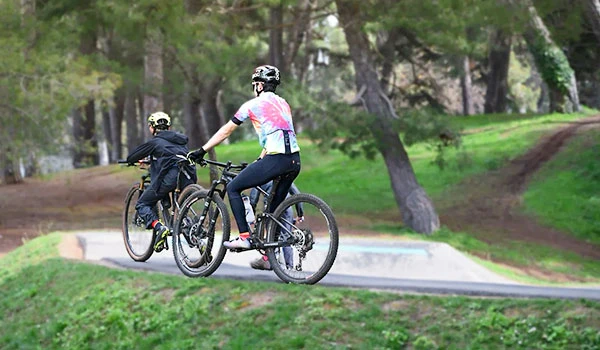 Duas pessoas pedalando de bicicleta em um parque com árvores e pista de terra, praticando mountain bike em um ambiente ao ar livre.