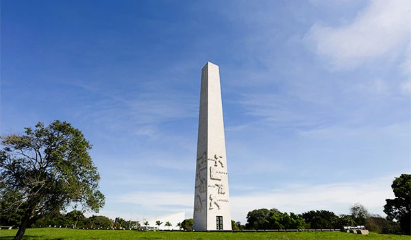 Imagem do Obelisco de São Paulo, no Parque Ibirapuera com árvores, céu azul e destaque para a estrutura alta e branca do monumento.