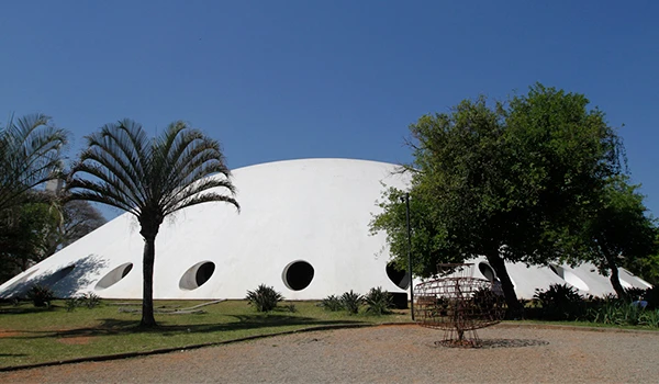 Estrutura arquitetônica moderna do Planetário no Parque Ibirapuera, com formato ovalada branca, cercada por árvores e um céu azul claro ao fundo, em um ambiente ao ar livre.