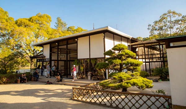 Imagem de um jardim zen com um chalé tradicional japonês rodeado por árvores verdes e pessoas apreciando o ambiente tranquilo ao ar livre no Parque Ibirapuera.
