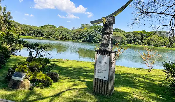 Esculturas em um parque ao lado de um rio, com árvores e céu azul ao fundo, destacando uma obra de arte moderna na natureza.
