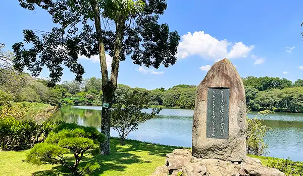 Paisagem de parque com lago, árvores e uma pedra com uma placa de inscrição ao lado da água, sob céu claro e nuvens.