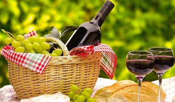 Cesta de piquenique com garrafas de vinho, uvas, pão e vinho tinto em um ambiente ao ar livre e ensolarado, perfeito para momentos de lazer ao ar livre.
