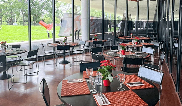 Ambiente de restaurante elegante com mesas decoradas com toalhas vermelhas, copos de vinho, flores e utensílios de mesa, paredes de vidro com vista para o exterior.
