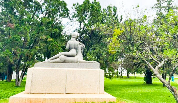 Escultura de uma mulher deitada, feita de material claro, em um parque com árvores verdes ao redor. Uma das obras de arte do Parque Ibirapuera.