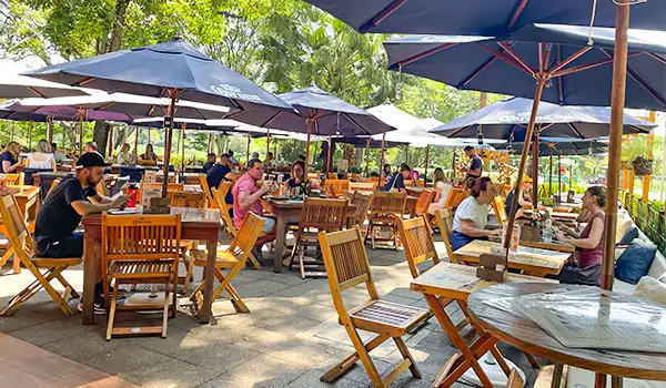 Varanda de restaurante ao ar livre com muitas mesas, cadeiras de madeira e guarda-sóis, rodeada por árvores verdes, clima agradável para refeições ao ar livre.