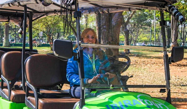 Mulher dirigindo um carrinho de golfe em um parque, rodeada de árvores, com expressão tranquila, usando jaqueta azul e óculos de proteção.