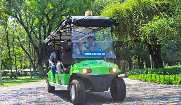 Imagem de um carrinho de golfe elétrico verde equipado para passeios turísticos em um parque, com duas pessoas a bordo e cercado por árvores verdes, o Ibira Circular.