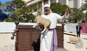 Um homem vestido em trajes tradicionais judaicos segura uma ovelha em frente a um altar. A imagem retrata uma cena bíblica que destaca rituais antigos.