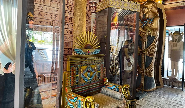 Imagem do interior de uma réplica de tumulo egípcio, apresentando artefatos como uma cadeira decorada e figuras de deuses egípcios.