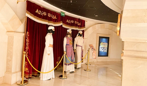 Maniquins vestindo trajes tradicionais, expostos em um ambiente elegante com cortinas vermelhas e um cenário histórico ao fundo.