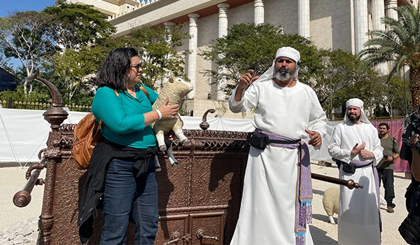 Cenário interativo com a participação de uma mulher segurando uma ovelha de pelúcia e homens vestidos com trajes tradicionais, promovendo a cultura e tradições vivas, cenas durante a visita guiada ao Templo de Salomão.