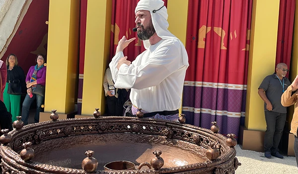 Guia vestindo roupas brancas e um turbante apresentando uma atividade interativa em um ambiente histórico com público ao fundo, durante a visita guiada ao Templo de Salomão.