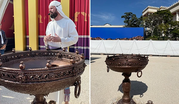 Imagem de um homem vestido com trajes tradicionais e um grande vaso decorativo em um ambiente ao ar livre. O vaso possui detalhes elaborados e está em um cenário festivo.