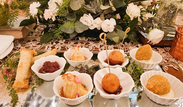 Delicioso prato variado com aperitivos finos dispostos artisticamente em pequenas tigelas brancas, ao lado de flores e uma toalha rendada.