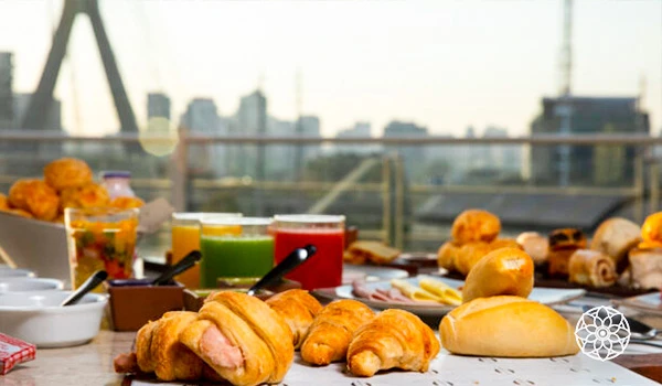 Delicioso café da manhã com croissants e pães variados em uma mesa com vista para a cidade. A refeição é acompanhada de sucos e frutas frescas.
