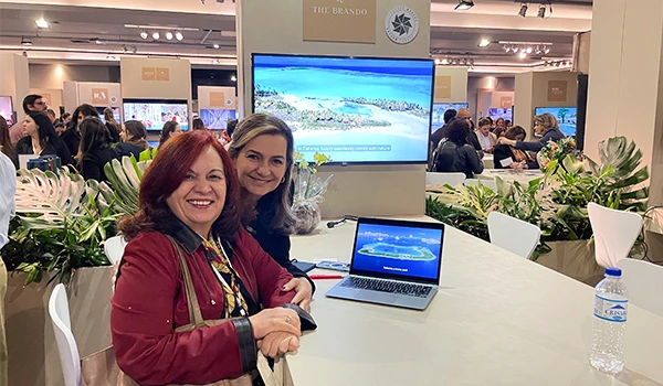 Deusa Rodrigues evento sobre casamentos em São Paulo, sentadas à mesa com um laptop exibindo uma imagem de praia ao fundo. O ambiente possui plantas e pessoas.