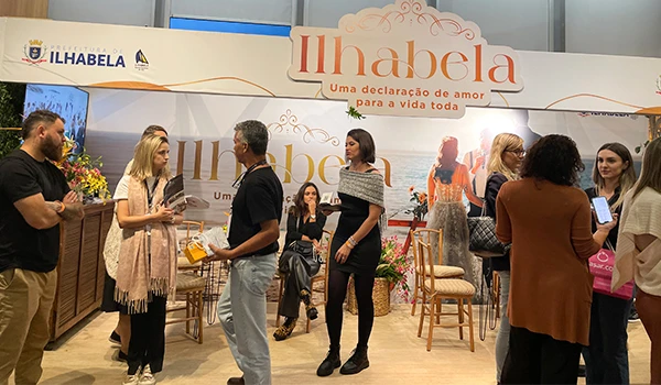 Evento sobre casamento em São Paulo, com estande de Ilhabela, com diversas pessoas interagindo ao fundo de uma decoração alusiva à cidade. Uma celebração de amor e cultura local.