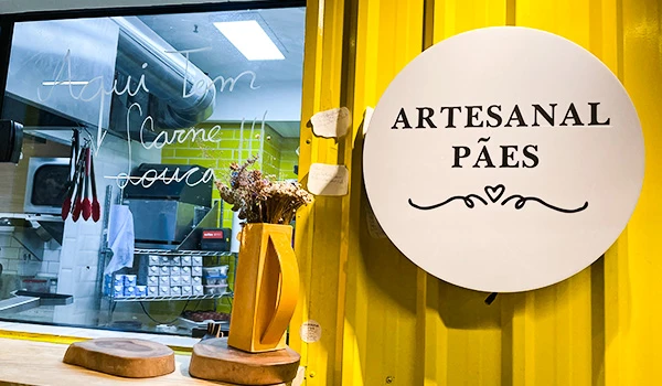 Interior de uma loja artesanal de pães, com um letreiro indicando 'Artesanal Pães' e uma decoração com áreas de trabalho visíveis ao fundo.