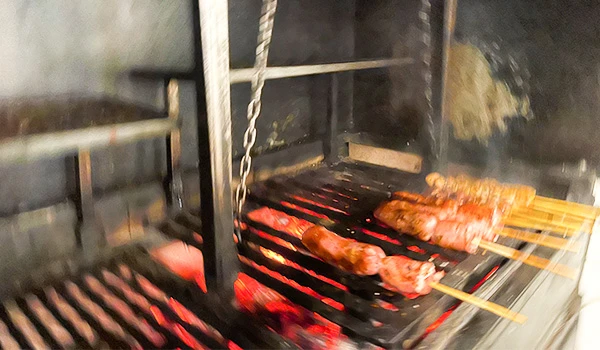 Churrasco de carne sendo assado em churrasqueira a carvão, destacando a brasa e os espetos de madeira com carne suculenta e dourada, ideal para churrasco brasileiro. Mercado de Santo Amaro restaurantes.