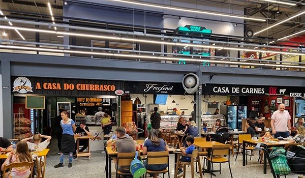 Imagens de uma churrascaria chamada 'A Casa do Churrasco' localizada dentro de um shopping. Clientes estão sentados nas mesas enquanto funcionários atendem e fazem pedidos. O ambiente é movimentado e confortável, ideal para quem aprecia churrasco de qualidade.